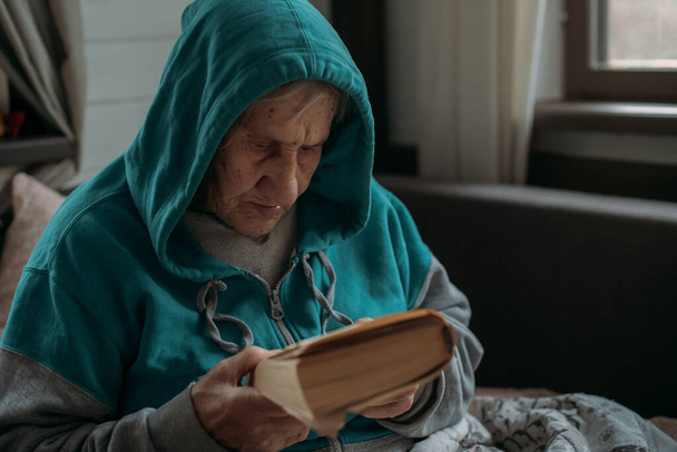 Mummi istuu sohvalla lukemassa kirjaa. Vanhempi nainen, eläkeläinen, lepää olohuoneessa päivän aikana. Kävelijät sängyn vieressä - Valokuva, kuva
