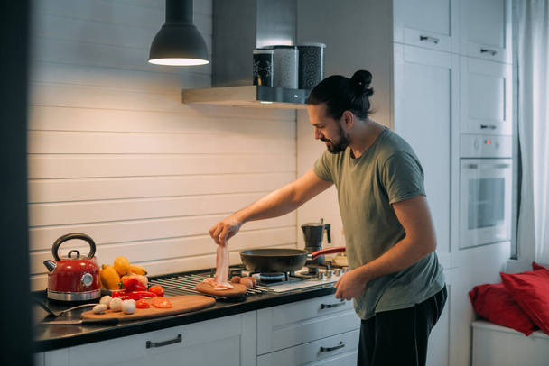 Een man bereidt ontbijt in de keuken. Jonge knappe blanke man bereidt eten voor zichzelf voor de lunch op een gasfornuis in een grote lichte keuken - Foto, afbeelding