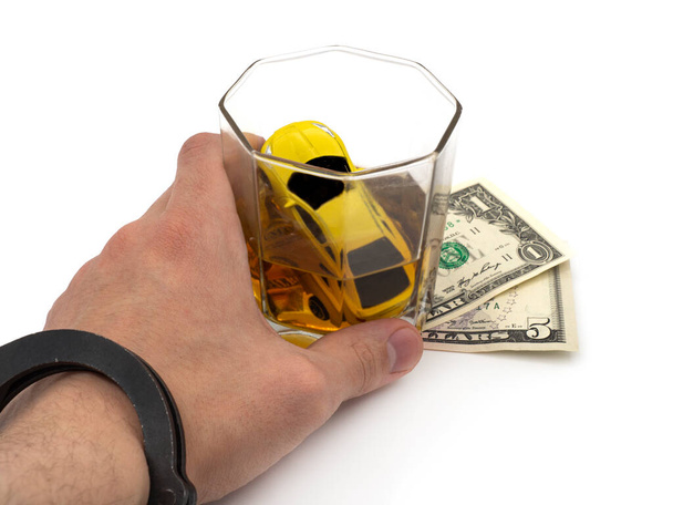 La mano de un hombre esposado sostiene un auto ahogándose en un vaso de whisky parado en billetes de dólar. El concepto de publicidad social, arresto por conducir ebrio. Estudio foto aislada sobre fondo blanco. - Foto, imagen
