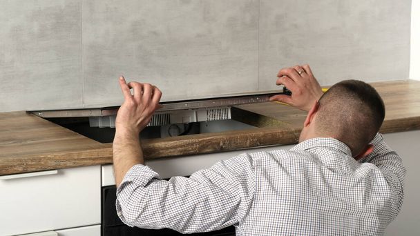 Ein junger Reparateur installiert ein schwarzes Induktionsherd in einer modernen weißen Küche im skandinavischen Stil mit Betonwand. Elektriker, mach 's selbst. Hausarbeit.  - Foto, Bild