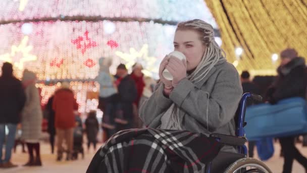 Молодая женщина с серыми дредами в инвалидной коляске на рождественском празднике на открытом воздухе - пить кофе из чашки - Кадры, видео