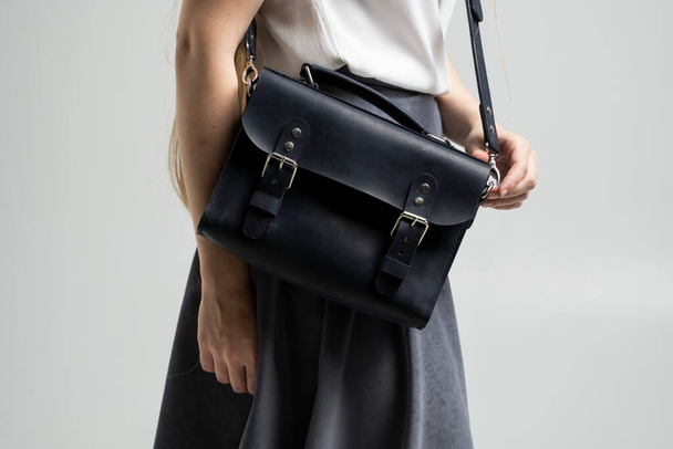 Small black leather bag over a woman shoulder on a white background. Shoulder handbag. Woman in a white shirt and grey skirt and with a black handbag. Style, retro, fashion, vintage and elegance. - Photo, Image