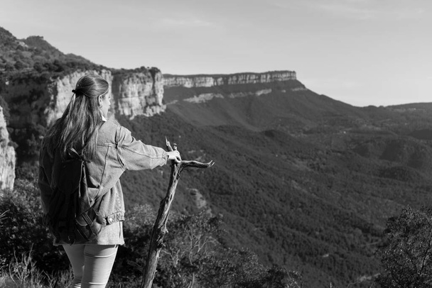 Beautiful young caucasian girl with small backpack looking mountains horizon while hold a tree branch.Travel freedom,exploring lifestyle concept.Black and white photo vintage retro style background. - Photo, Image