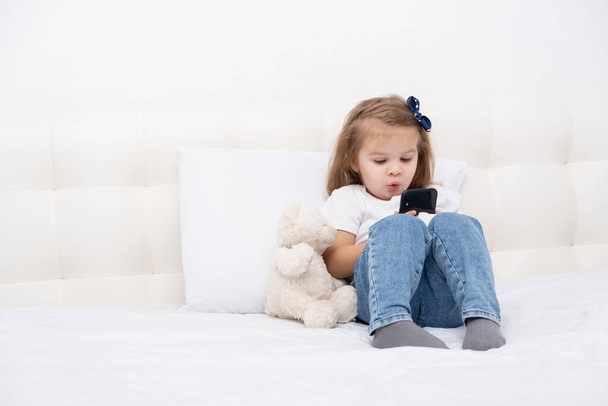 Μικρό κορίτσι με το χέρι στο cast κάθεται στο κρεβάτι χρησιμοποιώντας smartphone, βλέποντας κινούμενα σχέδια ή βίντεο εκπαίδευσης. - Φωτογραφία, εικόνα