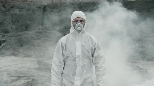 Technik laboratoryjny w masce i chemicznym kombinezonie ochronnym, spaceruje po suchym gruncie ze skrzynką narzędziową przez toksyczny dym - Materiał filmowy, wideo