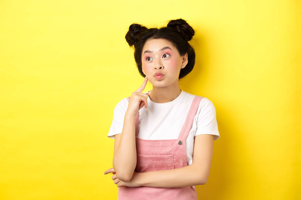 Summer lifestyle concept. Pensive glamour asian girl thinking, looking left at logo and dreaming, making choice, standing on yellow background - Foto, afbeelding