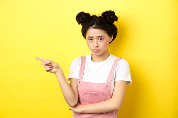 Sad and gloomy asian girl with glamour makeup, frowning unfair and pointing left at logo, looking disappointed, standing on yellow background - Photo, image