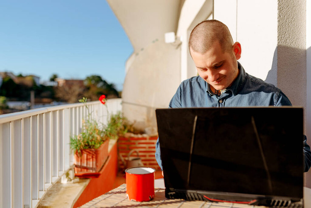 Mosolygó fiatalember borotvált fejjel használja a laptopját, hogy otthonról dolgozzon. az otthon kényelméből dolgozva. Vidám ember az interneten. - Fotó, kép