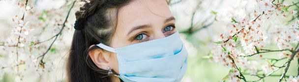花の木の間の公園に広がるコロナウイルスを避けるために、医療用フェイスマスクの若い女性. - 写真・画像