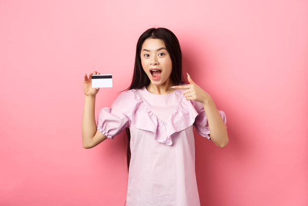 Happy cute asian woman showing plastic credit card, recommending bank special deal, standing against pink background - Foto, Imagen