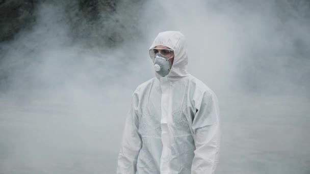 Laboratorioteknikko naamiossa ja kemiallisessa suojapuvussa kävelee kuivalla maalla myrkyllisen savun läpi - Materiaali, video