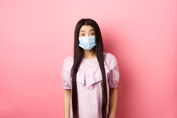 Covid-19, pandemic lifestyle concept. Surprised asian woman in medical mask look in awe, raising eyebrows with disbelief, standing against pink background - Foto, imagen