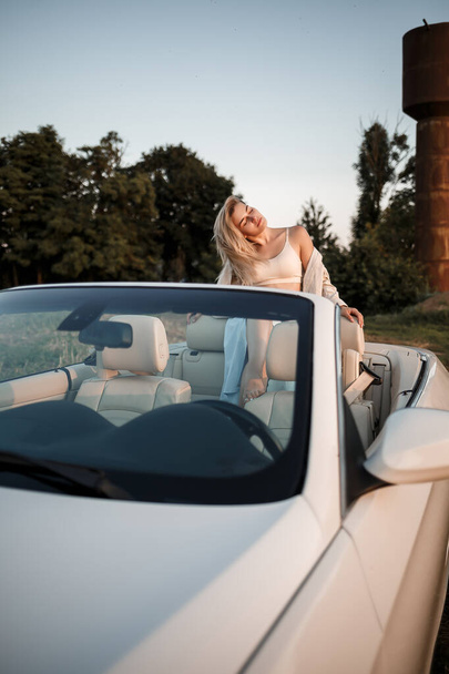 Een glamoureuze luxe meisje met blond haar glimlachend op een witte cabriolet. jonge succesvolle vrouw zitten in haar witte auto - Foto, afbeelding