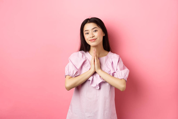 Cute azjatyckie dziewczyna powiedzieć dziękuję, uśmiechając się i patrząc szczęśliwy, pokazując namaste gest wdzięczności, stojąc w sukience na różowym tle - Zdjęcie, obraz