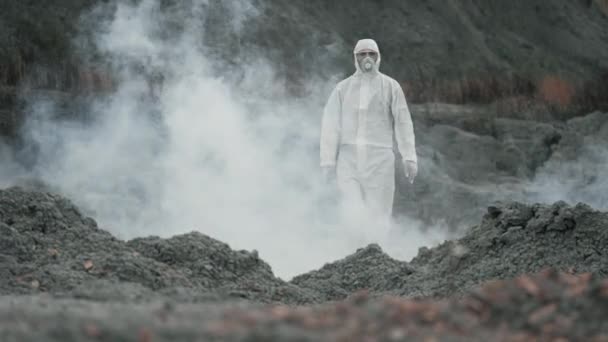 Técnico de laboratorio en una máscara y traje de protección química, camina sobre tierra seca con una caja de herramientas a través de humo tóxico - Imágenes, Vídeo