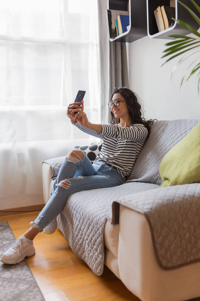 Jeune fille avec des lunettes assis dans son salon, elle prend un selfie avec son téléphone. - Photo, image