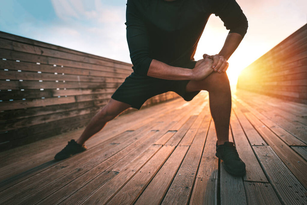Спортивный человек растягивает мышцы аддуктора ног и разогревается для тренировок на открытом воздухе на закате или на восходе солнца. Атлетик в черной спортивной одежде делает упражнения на растяжку. Спорт и здоровый образ жизни - Фото, изображение