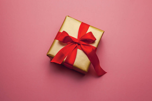 Holiday gift. Box in a gold gift wrapping with a red bow, on a pink background. Merry Christmas Valentines Day greetings. Postcard with love. - Fotoğraf, Görsel
