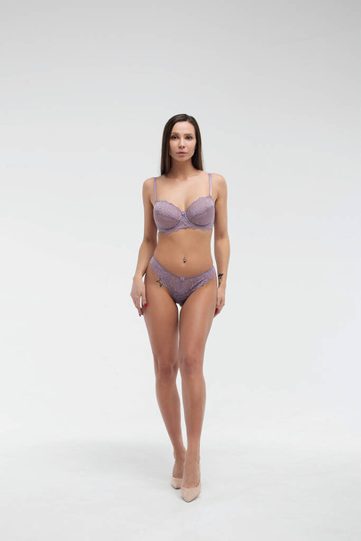 Coquette female model in lace purple bra and panties standing on white background in studio and touching hair while looking at camera - Φωτογραφία, εικόνα