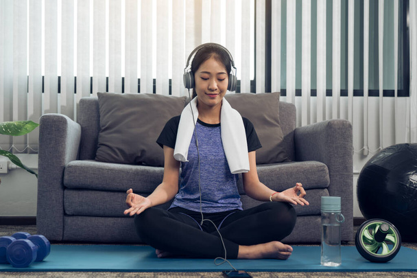 Mujer joven hermosa asiática usando teléfono inteligente y auriculares mientras medita y practica yoga en casa. - Foto, imagen