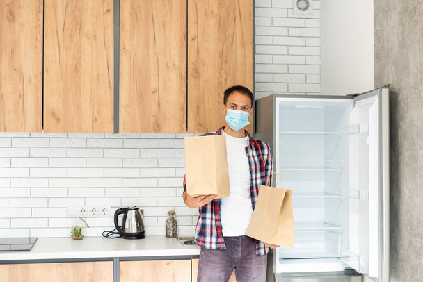 Ο διανομέας κρατάει μια χάρτινη σακούλα με φαγητό. Ο άνθρωπος που παραδίδει φαγητό σε χάρτινη σακούλα μιας χρήσης στον πελάτη στο σπίτι με μάσκα προσώπου. Παράδοση στο χρόνο του coronavirus - Φωτογραφία, εικόνα