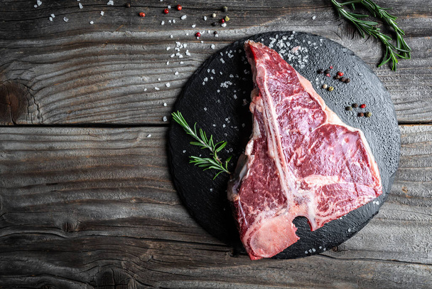Raw T-bone Steak or porterhouse steak for grill or BBQ with spices and herbs on slate board and woden rustic background, top view, border. - Φωτογραφία, εικόνα