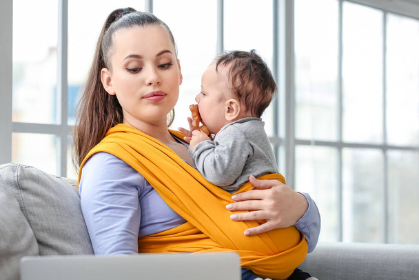 Jonge moeder met kleine baby in sling en laptop thuis - Foto, afbeelding