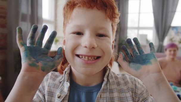 Portré felvétel vidám kisfiúról, amint festékes tenyereket mutat, a kamerába néz és mosolyog. - Felvétel, videó