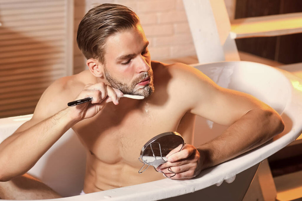 Man hygiene, health. Grooming man shave with razor look at mirror in bathtub. - Fotó, kép