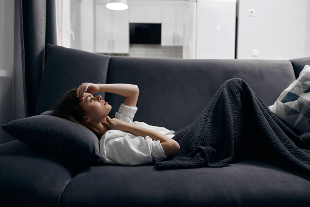 μια γυναίκα σε ένα άνετο δωμάτιο βρίσκεται σε έναν καναπέ που καλύπτεται με μια γκρι κουβέρτα - Φωτογραφία, εικόνα