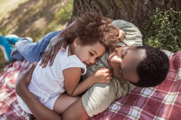 African american man hugging his cute little daughter and resting under the tree - Foto, immagini