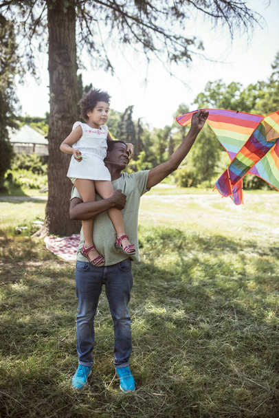 Dark-skinned man showing a kite to his daughter sitting on his shoulders - Foto, Bild