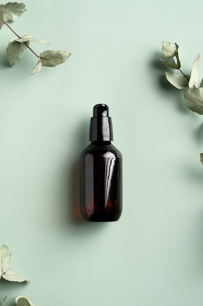 Dark amber glass bottle with serum and eucalyptus leaves on green background. Flat lay, top view. SPA natural organic cosmetics. - 写真・画像