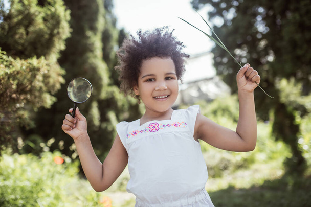 Curly-haired little girl holding a magnifier and looking cheerful - Fotografie, Obrázek