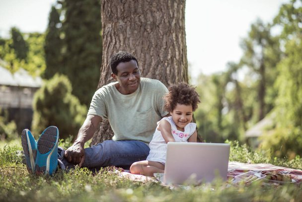 Curly-haired little girl sitting under the tree with her dad and watching something online - Photo, image