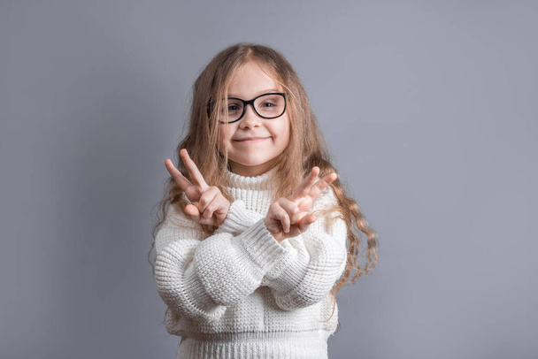 Портрет молодої привабливої маленької дівчинки з світлим волоссям в светрі, що показує v-знак, знак миру, жест перемоги на обох руках на сірому студійному фоні
 - Фото, зображення