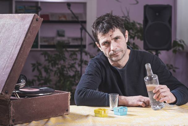 Un hombre solitario y triste se sienta en una mesa con un vaso de vodka en las manos y trata de encender un viejo gramófono. Salud mental. - Foto, Imagen