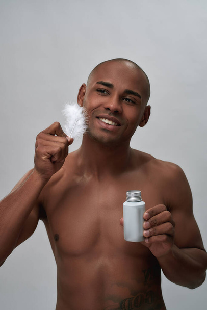 Shirtless handsome african american guy smiling, holding aftershave lotion, touching his facial skin with white feather, posing isolated over gray background - Foto, Bild