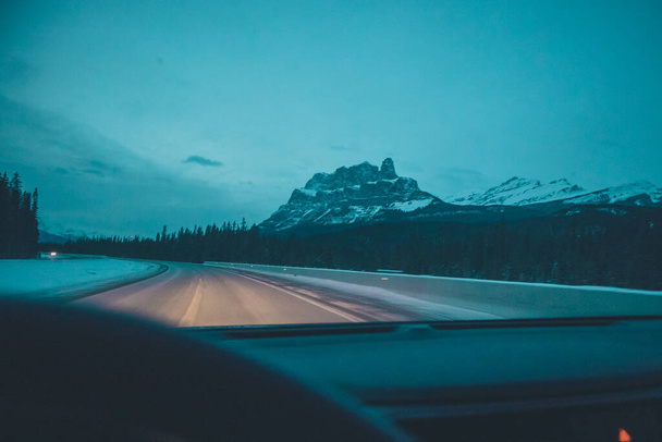 Driving with a car through empty road in canada with some snow and frost on the asphalt. Dangerous road situation, alone on the road, magnificent mountains nearby. - Foto, Imagem