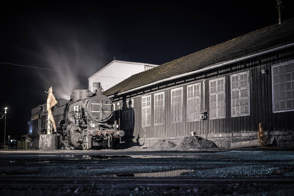 Stará parní lokomotiva se v noci nastartovala před kůlnou. Romantická fotka parního stroje v noci. Svislá fotografie - Fotografie, Obrázek