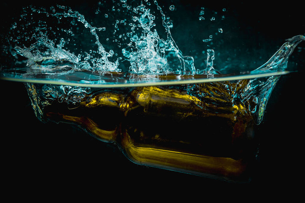 Brown beer bottle falls into water making big splash of drops and spray on a dark background. Juicy and fresh concept of refreshing drink in water. - Foto, Imagem