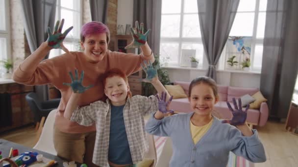 Happy Caucasian mother and little children looking at camera, making faces and showing palms with paint on them while making Easter decorations at home - Footage, Video