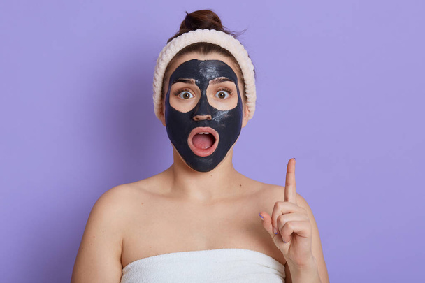 Astonished caucasian female with widely opened mouth pointing index finger up, having shocked facial expression, has great idea, lady with bare shoulders and hairband, doing skin care procedures. - Photo, image
