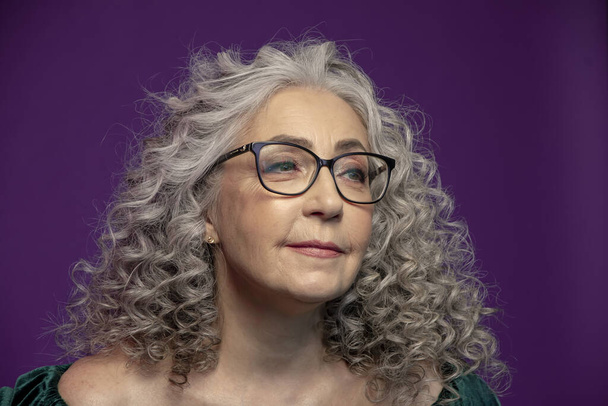 Studio muotokuva hymyilevästä iäkkäästä 60-65-vuotiaasta naisesta, jolla on lasit, harmaat kiharat pitkät hiukset, värillisellä taustalla, käsite: tyylikkäät mallieläkeläiset, aktiivinen elämä, - Valokuva, kuva
