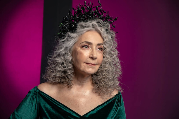Studioporträt einer älteren Frau von 60-65 Jahren in einem schönen Kleid mit einer Krone auf dem Kopf auf farbigem Hintergrund. Konzept: Stilvolle Rentner mit Model-Aussehen, aktives Leben. - Foto, Bild