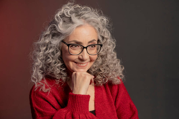 Studio πορτρέτο ενός χαμογελαστού ηλικιωμένη γυναίκα 60-65 ετών σε ένα κόκκινο πουλόβερ και γυαλιά, γκρι σγουρά μακριά μαλλιά, σε έγχρωμο φόντο. Έννοια: κομψοί συνταξιούχοι με εμφάνιση μοντέλου, δραστήρια ζωή. - Φωτογραφία, εικόνα
