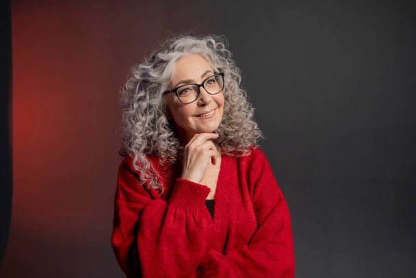 Studioporträt einer älteren Frau im Alter von 60-65 Jahren mit rotem Pullover und Brille, grauem lockigem, langem Haar auf farbigem Hintergrund. Konzept: Stilvolle Rentner mit Model-Aussehen, aktives Leben. - Foto, Bild