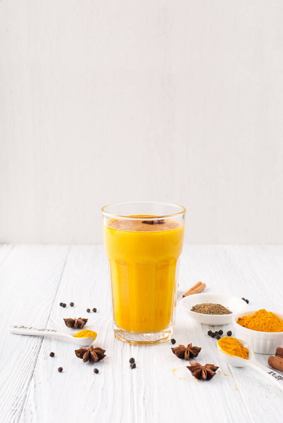 Golden turmeric latte in glass, spices and cinnamon on white wooden background. Selective focus, copy space. Trendy Asian natural detox beverage with spices for vegans - Foto, Bild