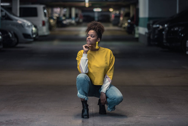 Full body of confident contemplative young African American female in bright yellow shirt and stylish earrings squatting and looking away pensively in underground parking - Photo, Image