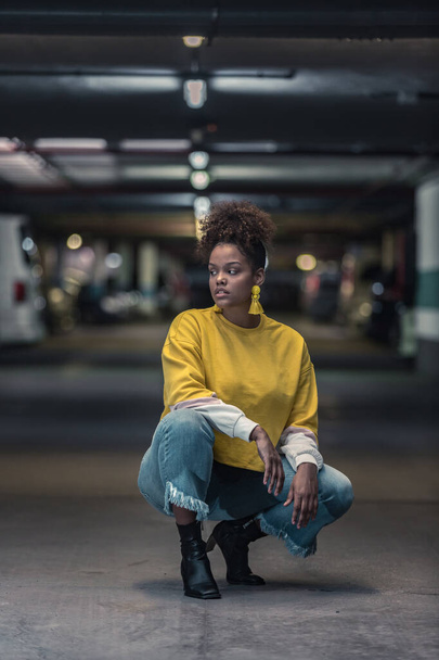 Full body of serious millennial African American female in modern trendy yellow shirt and jeans hunkering down and looking away in underground parking lot - Photo, Image